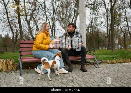 Young happy couple sitting on red bench in autumn park with cute Jack Russell dog, holding coffee cups, smiling and interacting playfully in cozy casu Stock Photo