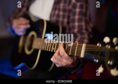 close-up horizontal image showing the hands of a male person playing an acoustic guitar. The person wears a red and dark plaid shirt. Stock Photo