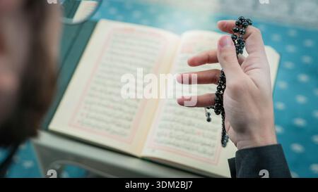 Close up of Muslim hands holding prayer beads over the Holy Quran. Misbaha and Quranic verses during Islamic worship. Ramadan spiritual reflection and Stock Photo
