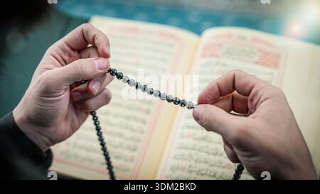 Close up of Muslim hands holding prayer beads over the Holy Quran. Misbaha and Quranic verses during Islamic worship. Ramadan spiritual reflection and Stock Photo