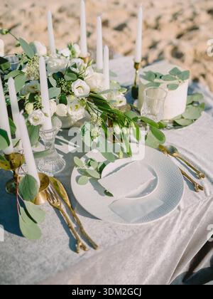 Elegant outdoor wedding table with white flowers, candles and cake on sandy beach, romantic luxury celebration setting. Stock Photo