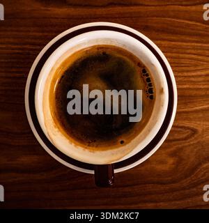 Top-down view of a fresh espresso in a white porcelain cup with thick crema bubbles and foam on a saucer, placed on warm wooden table texture Stock Photo