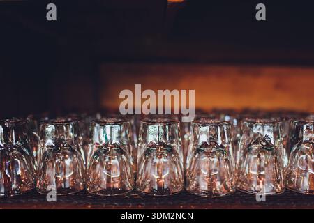 Rows of inverted glassware on bar shelf, warm low light, wooden counter, shallow depth of field, sparkling reflections, ready for bartender service Stock Photo