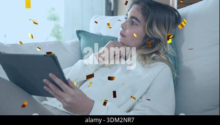Reclining woman using tablet, shirt on sofa with teal pillow, gold confetti, sheer curtains, window Stock Photo