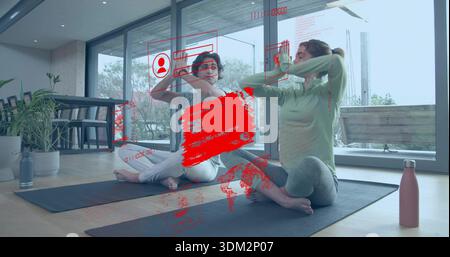 Stretching two women friends wearing leggings and tops in living room, with yoga mats, bottles, HUD Stock Photo