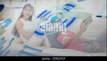 Smiling woman watching reclining friend in red shirt holding phone on sofa at home, HUD overlaying Stock Photo