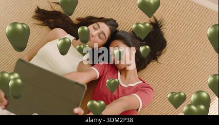 Lying friends taking selfie on woven rug with tablet, white top and red tee, green hearts Stock Photo