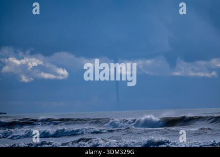 A dramatic weather event showing a waterspout funnel cloud descending from dark storm clouds over a turbulent ocean with crashing waves. Stock Photo