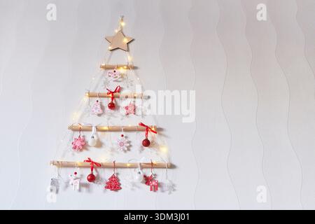 Creative hanging wall Christmas tree made of branches, decorated with red and white wooden toys and warm LED fairy lights on a light background. Stock Photo