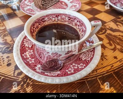 Vintage porcelain cup with red floral patterns filled with dark aromatic coffee, served on a matching saucer with a silver spoon on a decorative Stock Photo