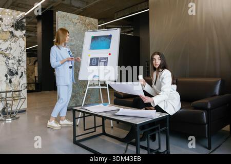 Professional women collaborating, one presenting information on a whiteboard, the other reviewing documents during an important team meeting Stock Photo