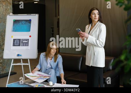 Two young women collaborating on a business project, analyzing charts and discussing data in a modern office environment Stock Photo