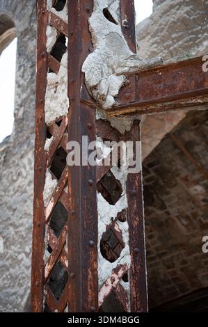 Close-up of a rusted metallic lattice column and beam junction in an abandoned factory, showing exposed rivets, heavy corrosion, and broken cement coa Stock Photo