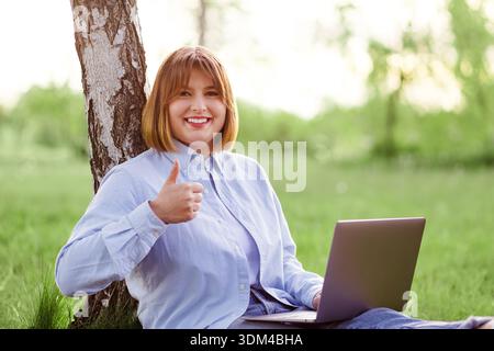 Photo portrait of pretty young woman smiling teeth sitting in green park working on laptop smiling Stock Photo