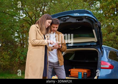 Man and woman using smartphone near open car trunk with suitcase in autumn forest. Travel and route planning Stock Photo