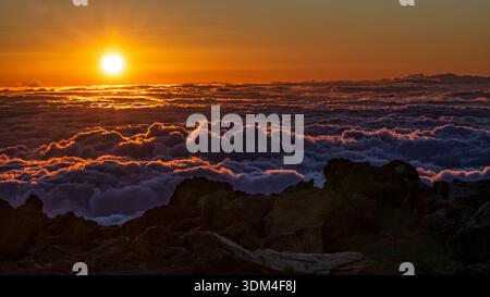 The sun sets above a roiling cloudbank, seen from the Haleakalā Summit at golden hour, In Haleakalā National Park, Hawaii. Stock Photo