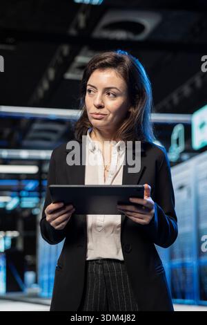 Admin in data center using tablet to inspect infrastructure rackmounts, updating network systems. IT specialist using device to ensure stable server farm operations, overseeing equipment Stock Photo