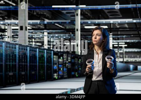 Server room manager using VR equipment to do maintenance on storage infrastructure rigs. Female engineer uses virtual reality gear to inspect high tech data center electronics Stock Photo