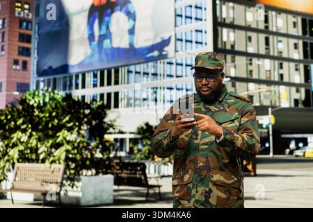 Adult male soldier in uniform navigates mobile screen on downtown street, showing disciplined confidence and respectful presence as he checks online page near urban buildings. Stock Photo