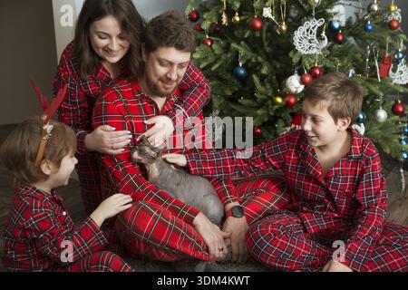 family mom, dad and two children brothers on Christmas morning at home. dad and kids stroke their cat sitting near the Christmas tree. Christmas presents || Model released Stock Photo