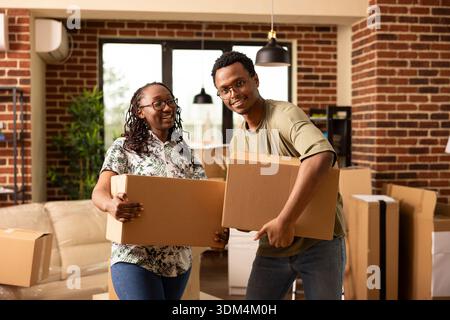 Young african american couple stands in modern living room, holding packed boxes while moving into new apartment. Portrait of boyfriend and girlfriend carrying belongings together on moving day. Stock Photo
