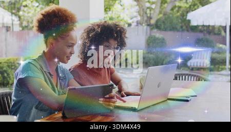 Collaborating two mid adult women leaning over laptop and tablet on sunny backyard patio, with mug Stock Photo