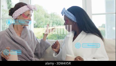 Chatting two women in robes headbands eye patches, holding mugs on sofa showing HUD overlays Stock Photo