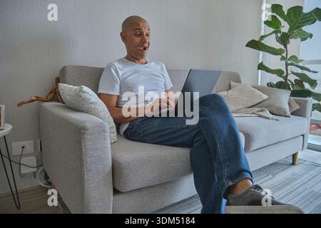 Middle-aged man sitting on sofa, reacting with surprise to content on his laptop screen Stock Photo