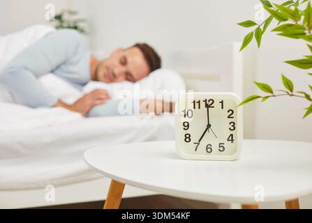 Alarm clock showing 7 AM on table with peaceful man sleeping comfortably under blanket on background Stock Photo
