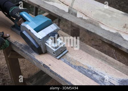 Focused carpenter at work, using an electric planer for woodworking. This skilled craft of carpentry involves smoothing wooden plank for construction Stock Photo