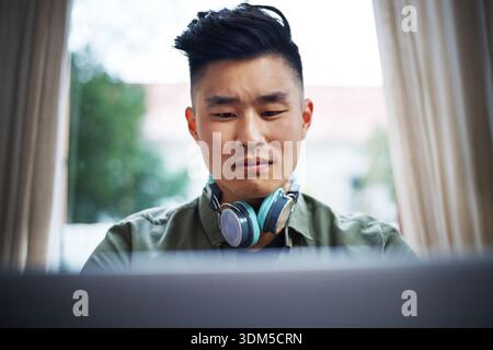 Headphones, programmer or asian man with laptop for remote work, system maintenance or development. Tech, freelancer or developer in home for software Stock Photo
