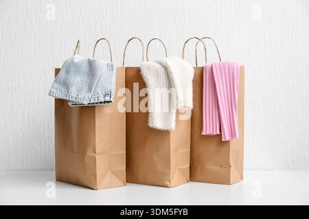 Paper shopping bags with different sweaters and jeans on table against white wall Stock Photo