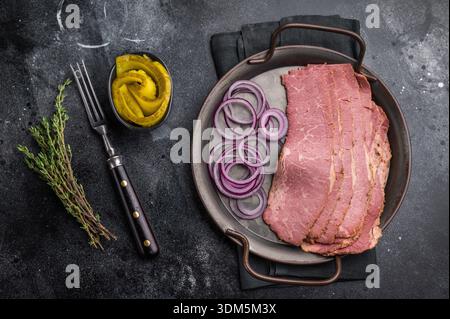 Thinly sliced pastrami on a metal plate with red onion rings and pickled cucumbers, placed on a textured dark stone background. Smoked deli beef ready Stock Photo
