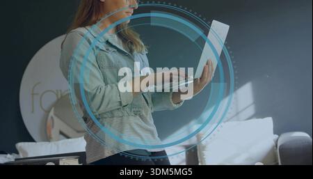 Typing mid-adult woman wearing denim jacket, striped shirt, holding laptop in living room, with HUD Stock Photo