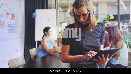 Presenting woman in glasses wearing dark ribbed top guiding team at conference table with tablet Stock Photo