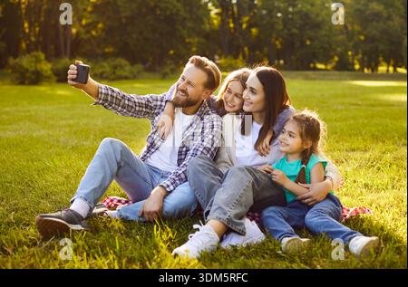 Happy parents with two children sitting on grass in sunny green park and taking selfie together. Stock Photo