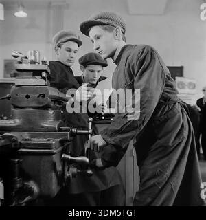 Slaviansk, Donetsk region, Ukrainian SSR, USSR. 1970s. An authentic archival genre scene of vocational training in a railway depot workshop. A young apprentice in a work apron and a cap operates a lathe machine, carefully turning a metal part. In the background, two other students or young workers are discussing a technical drawing or a blueprint. The image captures the process of learning a trade, the combination of theory and practice, teamwork and the industrial atmosphere in the peaceful Donbass. Stock Photo