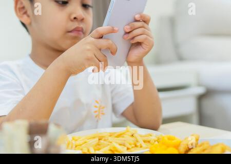 Selective focus of Asian child using mobile phone while eating some snack at home. Kids addicted phone. Stock Photo