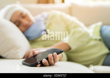 Selective focus of mobile phone on mature tired female with cancer sleeping laying relax on bed. Cancer or disease concept. Stock Photo