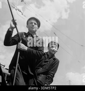 Slaviansk, Donetsk region, Ukrainian SSR, USSR. 1970s. An expressive archival low-angle portrait of two railway workers standing on a locomotive or a maintenance vehicle. The men are wearing vintage dark uniforms and peaked caps with emblems. One worker holds a metal handrail while looking forward with his colleague against the background of the cloudy sky and overhead catenary wires. The image captures the professional focus, the aesthetics of the transport profession and the sunny atmosphere in the peaceful Donbass. Stock Photo