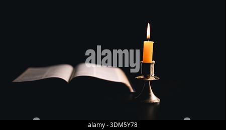 Dark, an antique candle cast its light on the old Bible, its flames flickering gently over the book on the table, symbolizing the enduring essence of Stock Photo