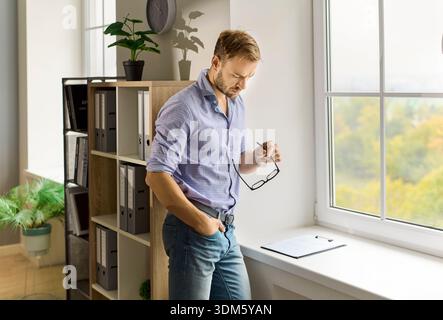 Thoughtful man by window in office room with glasses, reflecting on plans Stock Photo