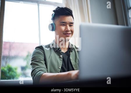 Creative, man and laptop in home with headphones, music and remote work for video editing project. Asian person, smile and audio tech in house with Stock Photo