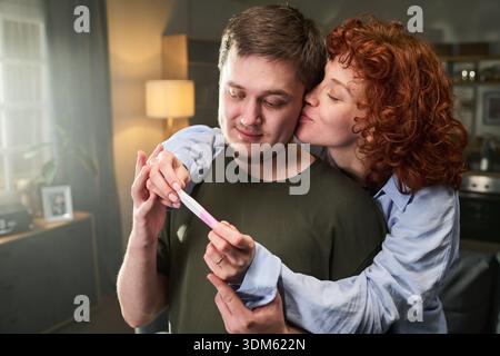 Caucasian young adult woman embracing and kissing Caucasian young adult man from behind while both holding pregnancy test, man looking down with thoughtful expression Stock Photo