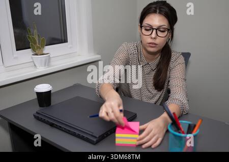 A woman sits at a desk in an office. She writes on a colorful sticky note with a pen. A laptop sits closed next to her. A plant and a cup of coffee are on the desk as well. Stock Photo