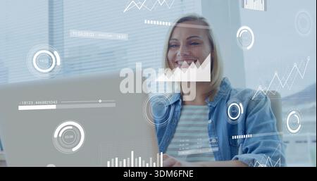 Smiling woman working at desk in office, wearing denim jacket over striped top, viewing laptop data Stock Photo
