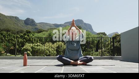 Sitting woman in blue top and leggings practicing yoga on terrace on mat with pink bottle Stock Photo