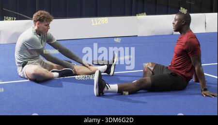 Stretching two male athletes in red and gray tops on blue turf at gym, wristwatch overlay Stock Photo