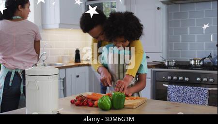 Woman guiding child's hands cutting vegetables in kitchen, wearing aprons, with peppers and carrots Stock Photo