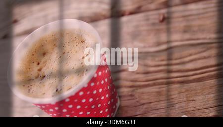 Sitting red polkadot cup on wood table with frothy coffee, shadow lines and sunlight, copy space Stock Photo